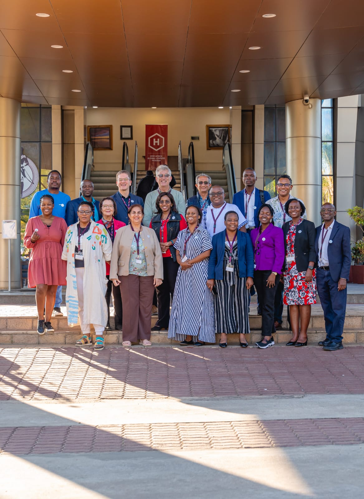 PCAT partners from eleven countries pose together on the entrance steps of the meeting venue in Lusaka, wearing lanyards and PCAT logo badges.